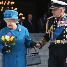 Queen Elizabeth wears a blue coat and matching hat and carries a bunch of tulips while her husband Prince Philip holds her hand
