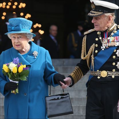 Queen Elizabeth wears a blue coat and matching hat and carries a bunch of tulips while her husband Prince Philip holds her hand