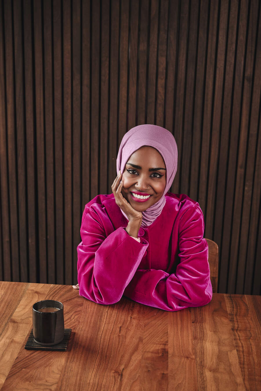 Nadiya Hussain sitting at a wooden table and smiling at the camera