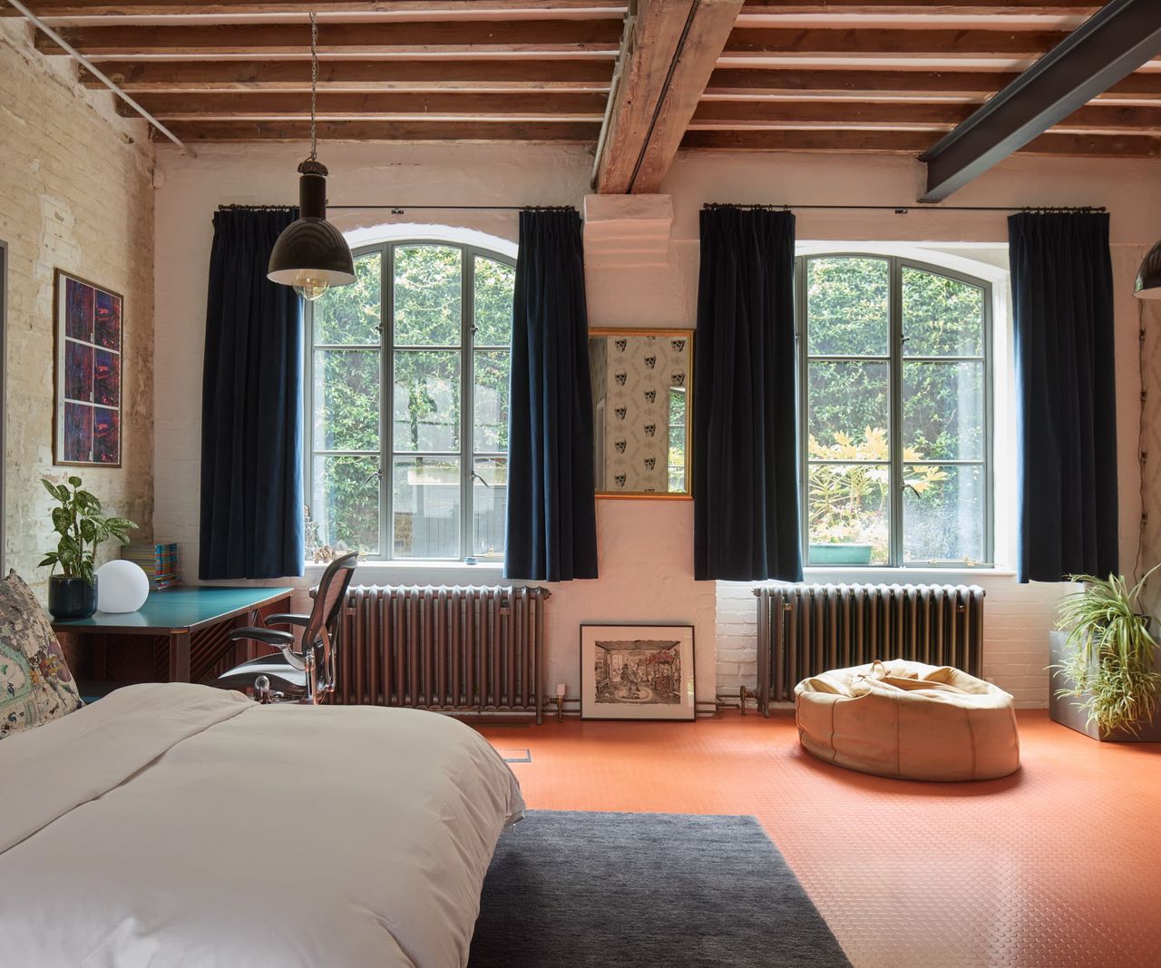 A bedroom with a wooden beams above and an office space next to the bed