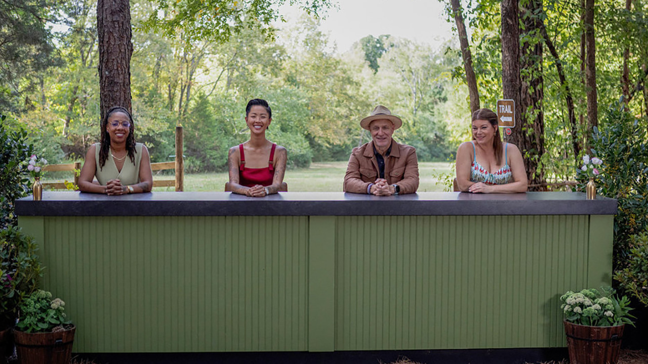 Kristen Kish sits with the other Top Chef judges during an outside challenge on Top Chef. They are at a green desk. 
