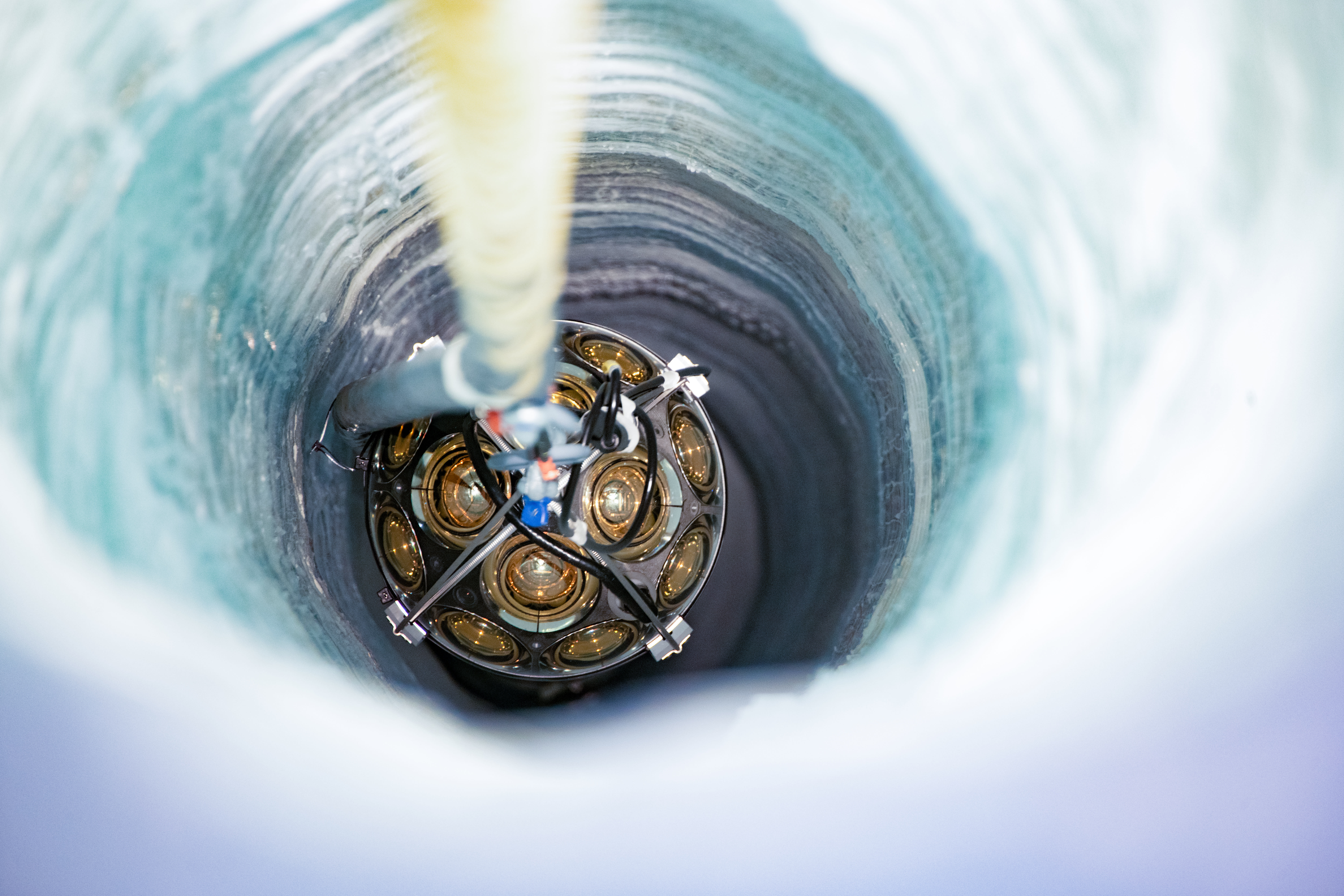 A top-down view of a detector being lowered into an icy white tunnel from a hook, its gold metal pieces glowing in the light.