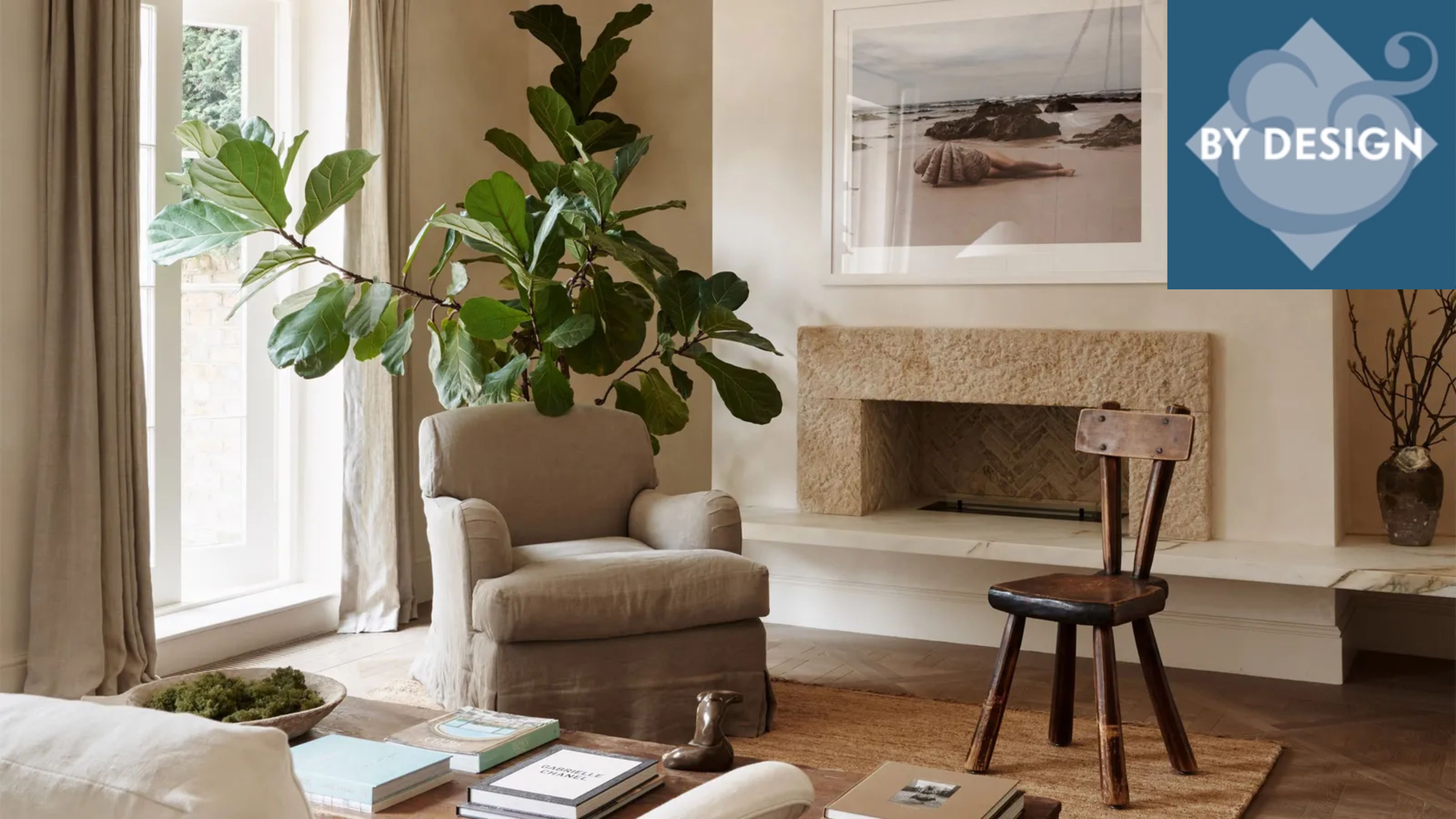 Cozy neutral, minimalist living room. Armchair and wooden chair in front of fireplace, greenery and artwork in background, back of sofa and coffee table in foreground
