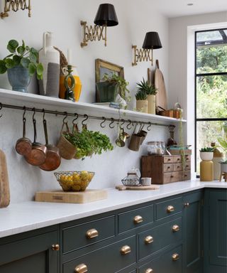 A charming shaker-style kitchen with deep green cabinetry accented by brass cup handles. A white marble countertop contrasts with the cabinetry, extending along the length of the kitchen.