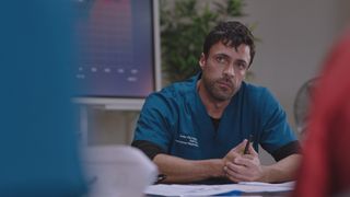 Flynn sitting at his office desk looking pensive while looking at out of shot colleagues. 