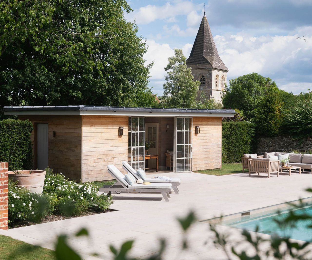 A timber-clad flat-roof pool house with Crittall-style glazed doors and sun loungers on a limestone terrace beside a pool, with a church tower visible beyond.