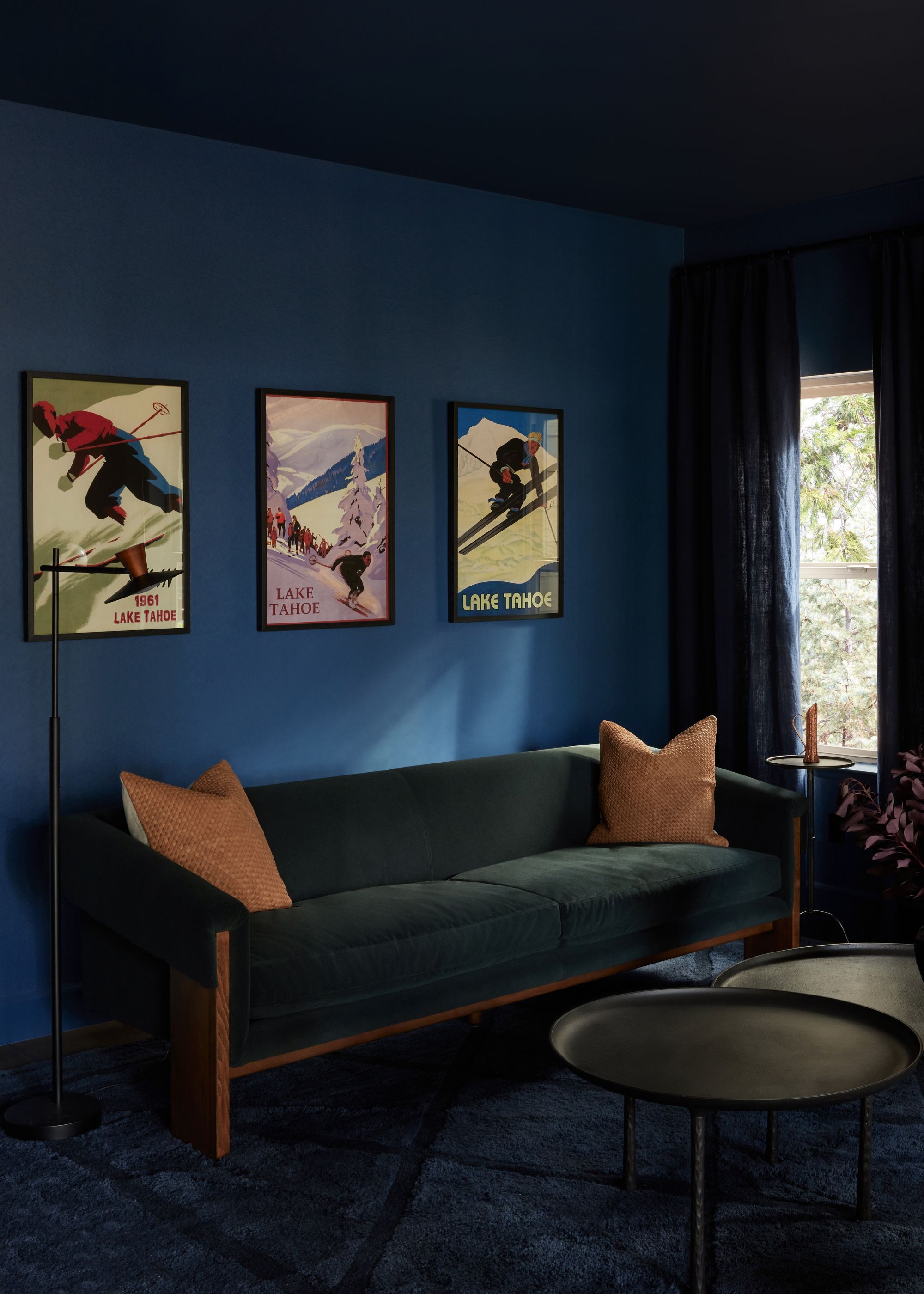 A snug room, color-drenched dark blue with a dark green sofa with orange cushions, a dark blue carpet, and three artworks on the wall.