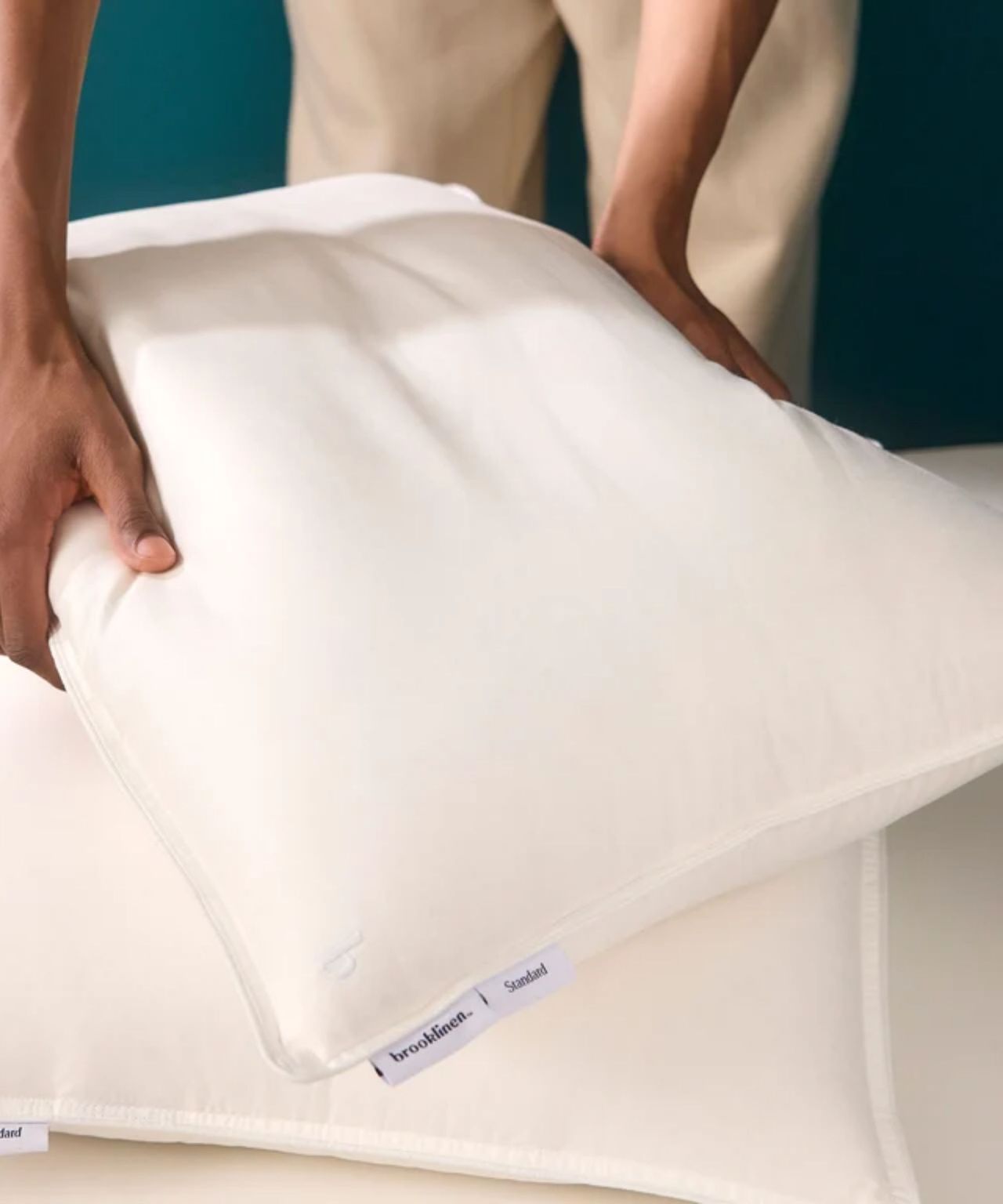 Two hands stacking two Brooklinen down pillows against a teal wall