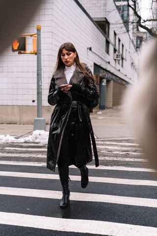 A woman is seen walking at New York Fashion Week street style wearing a black faux leather trench coat, white turtleneck, and black boots