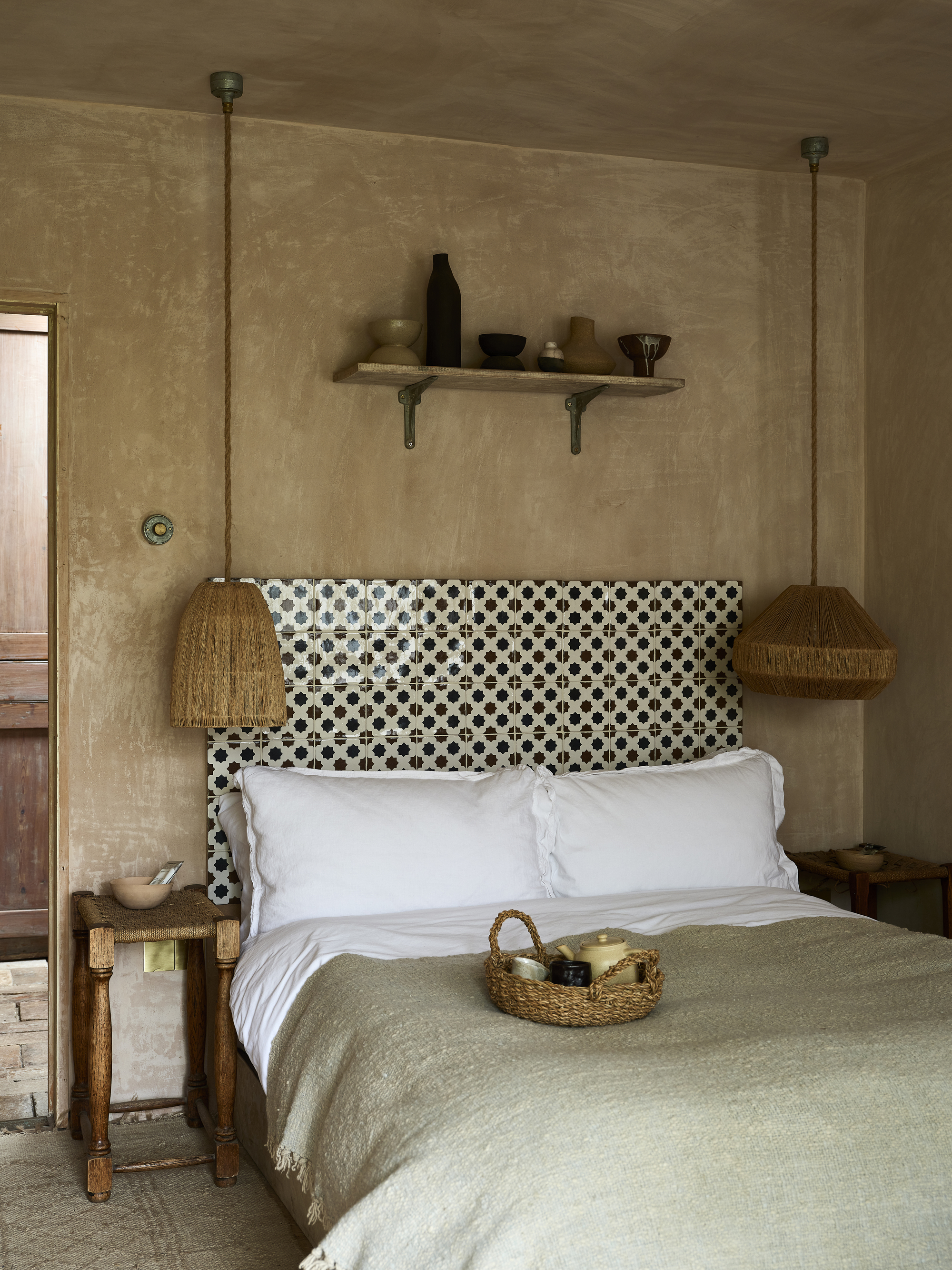 Image of a bedroom painted in a dusty pink limewash. There is a bed with white and sage green bedding and a tiled headboard made with black and white tiles. There are two rattan pendant lights hanging over wooden side tables on either side of the bed.