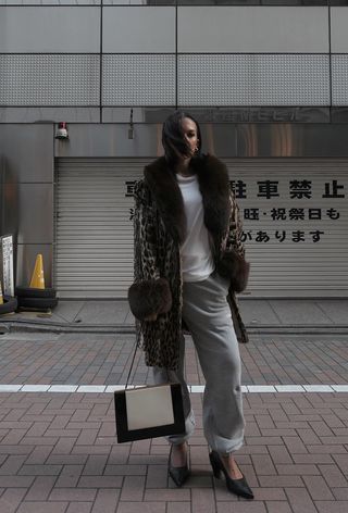 A winter travel outfit is shown in a photo of a woman standing outside wearing a leopard fur coat over a baggy white t-shirt styled with gray sweatpants, black pumps, and a black and white frame handbag