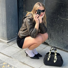 Hannah Juneva holding a black Coach bag wearing black sunglasses, a leather bomber, black shorts, white socks, and black pointed toe mules.