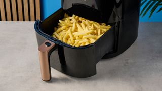 a photograph of yellow uncooked frozen french fries inside the basket of a large black 5.3 quart capacity air fryer with black touchscreen and rose gold embellishments sits upon a brown table with a blue background