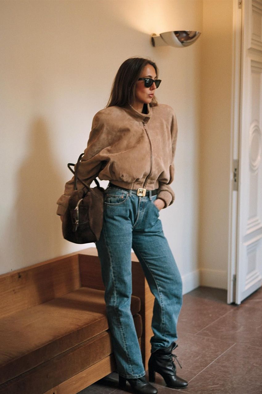 A woman standing inside a hallway wearing an '80s-inspired light tan suede bomber jacket styled with light-wash jeans, a black belt, black ankle boots, a dark brown suede bowler bag, and black sunglasses.