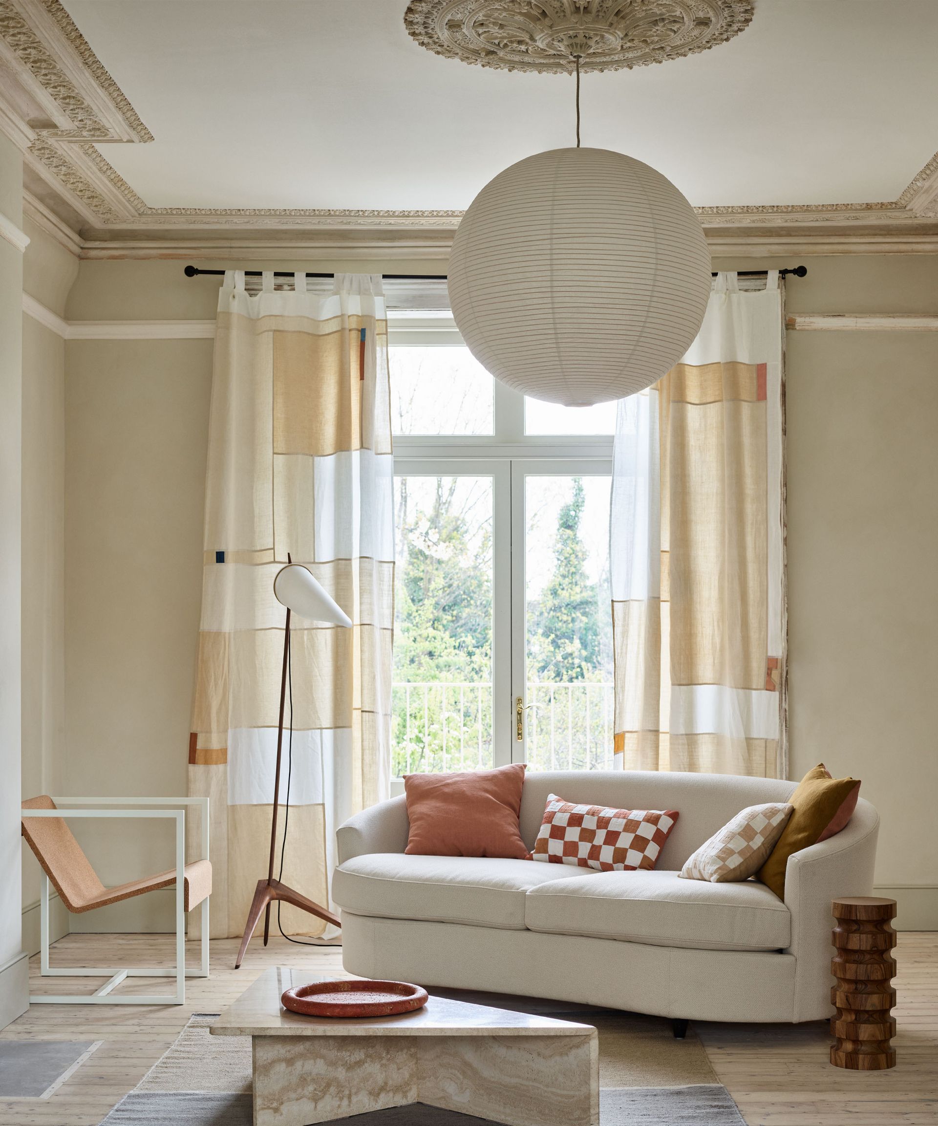 Calming, neutral modern living room with abstract square patterned curtains, light wooden flooring, rounded paper lantern shade, curved cream sofa, floor lamp, leather lounge chair, marble coffee table on rug