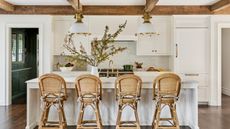 A white kitchen with wooden ceiling beams, rattan bar stools, and fall foliage displayed in a vase on the island