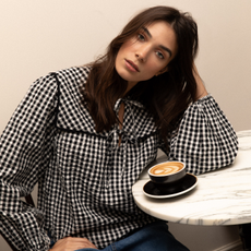 Model wearing a black and white gingham blouse sold at nobody's child with her arm on a cafe table and a coffee in front of her