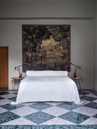 Modern bedroom with white walls and a tapestry hanging over the bed. The floors are a lavender and aquamarine checkerboard print made of terrazzo tile.