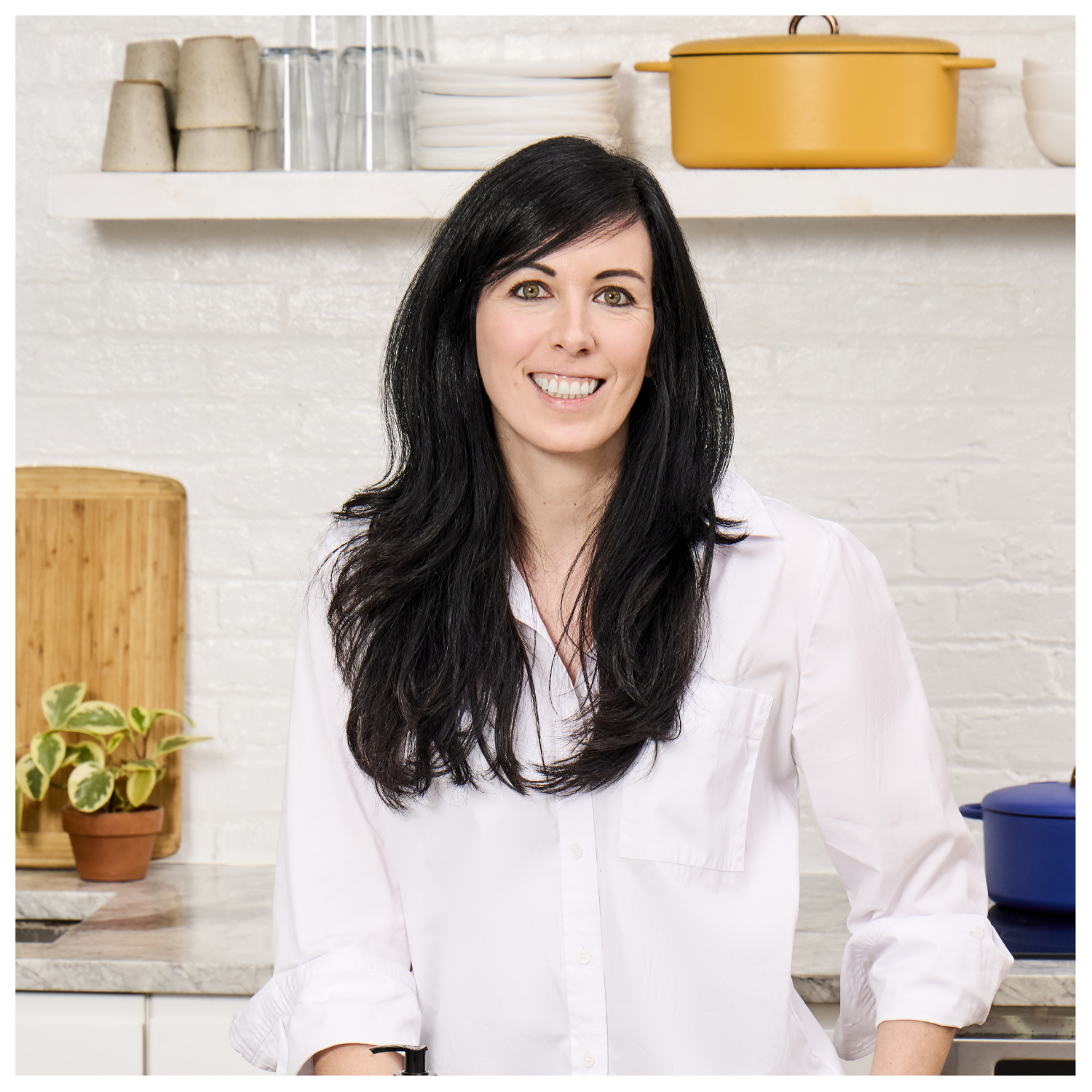 A headshot of a woman with long, shiny black hair in a white shirt, smiling in a kitchen