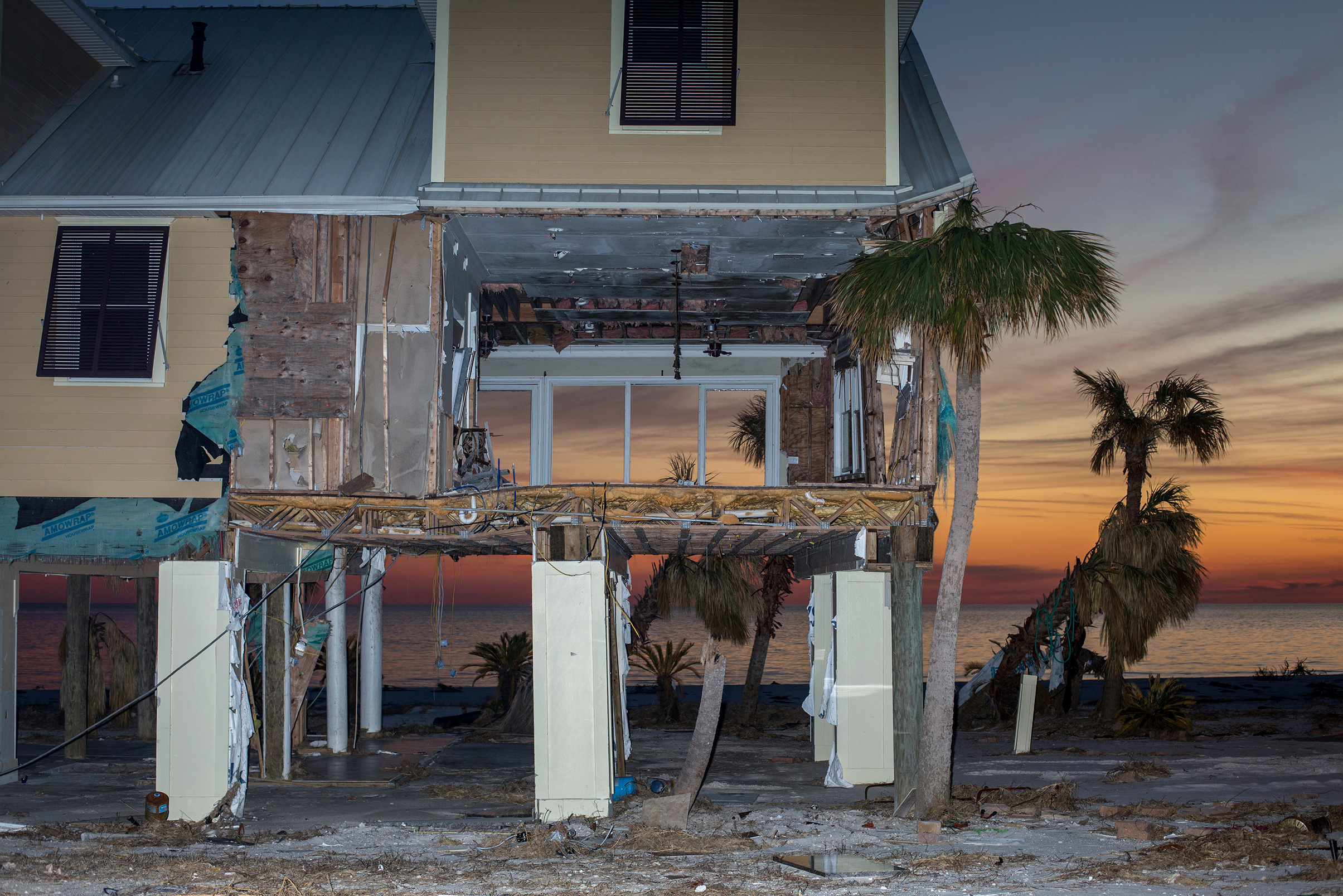 Partially destroyed elevated beach house with exposed interior and palm trees at sunset by the ocean