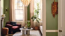 A cozy apartment living room with green painted walls. A red armchair in the far corner, a small round marble coffee table in front of it, and a cream rug.