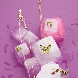 The Zuri Glass Butterfly Pendant Necklace puctured on a vibrant purple background surrounded by melting ice cubes