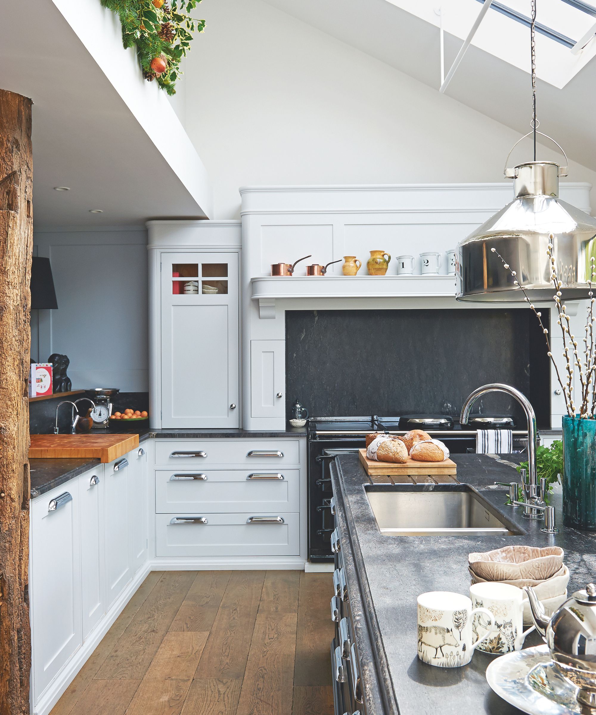 A festive decorated kitchen with white cabinets, wooden flooring, a large kitchen island with black marble countertops with a silver sink and faucet, and an exposed wood beam to the left. Above the beam is a shelf with festive foliage, and to the right is a hanging light with large silver lampshade.