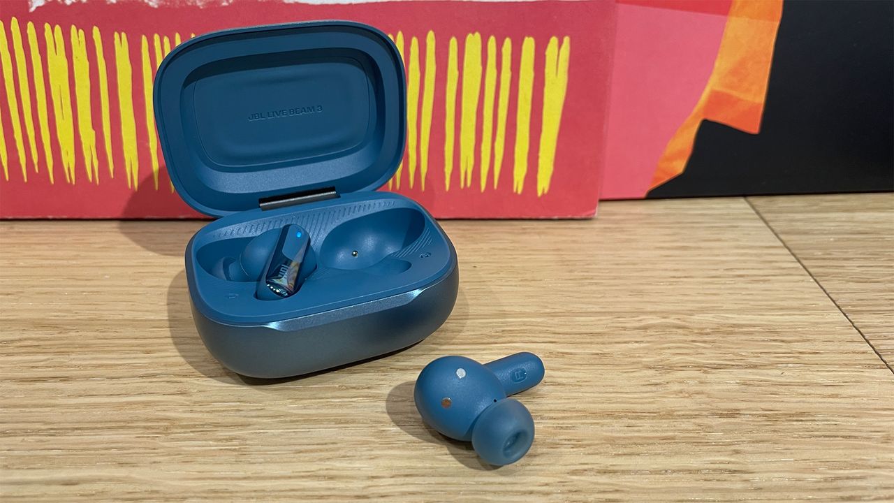 The blue case for a pair of JBL Live Beam 3 earbuds. The lid is open and one of the buds has been removed and placed on the wooden surface in front of the case.
