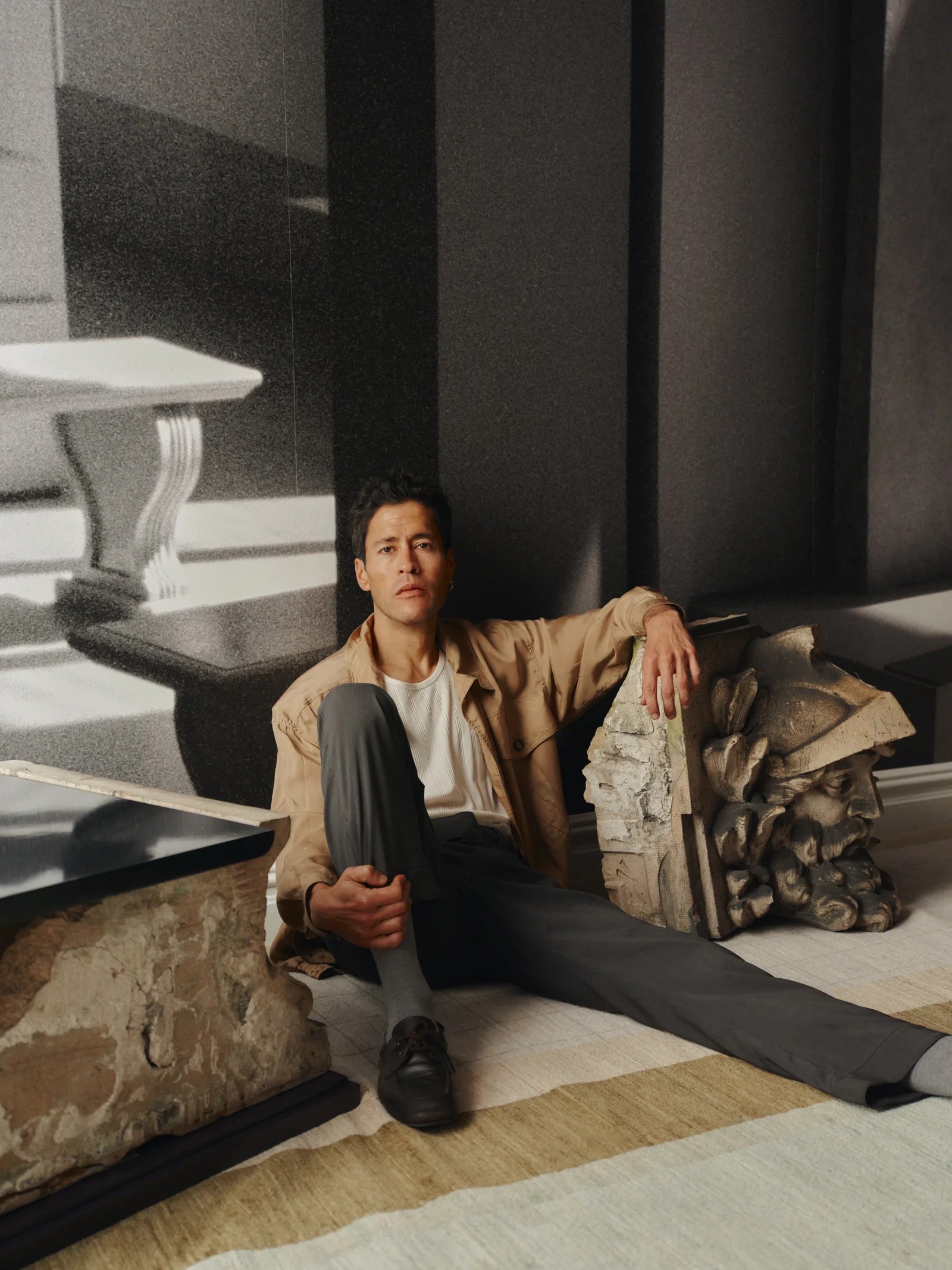 A man dressed in a sand-toned jacket over a white T-shirt, gray trousers, and loafers sits on a striped rug while leaning against a fragment of a marble capitel depicting a bearded soldier.