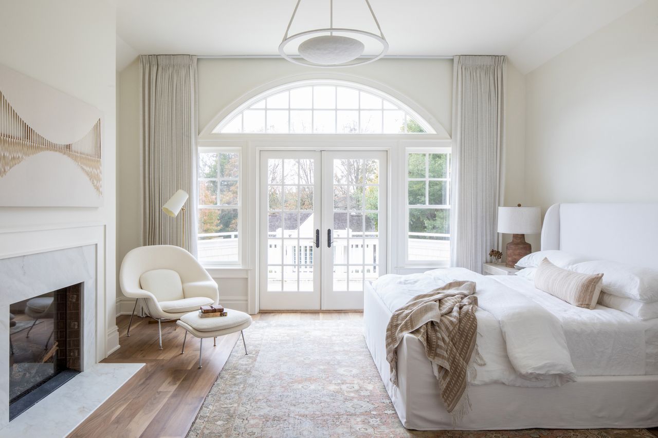 white bedroom with boucle chair and footstool, fireplace, modern art, antique rug, wooden floor, view outside 