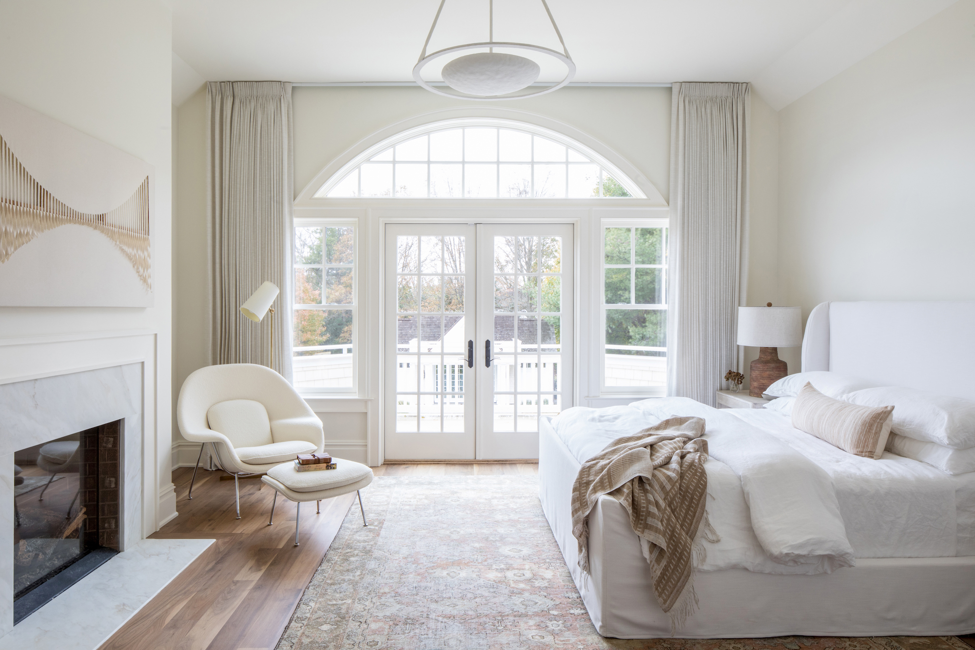 white bedroom with boucle chair and footstool, fireplace, modern art, antique rug, wooden floor, view outside