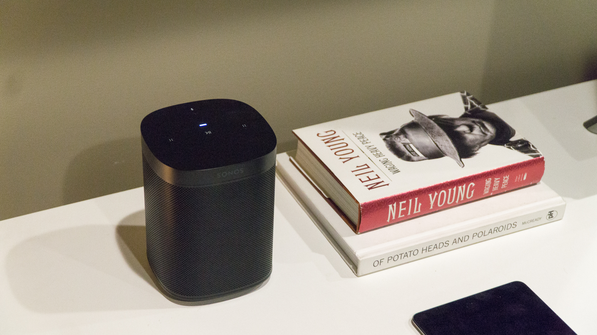 The Sonos One smart speaker in black pictured on a white surface next to a small stack of books.