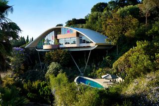 Garcia House A double storey house built on a hill with metal beams holding it up, a curved roof, large windows, trees all around it and a pool below it.