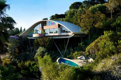 Garcia House A double storey house built on a hill with metal beams holding it up, a curved roof, large windows, trees all around it and a pool below it.