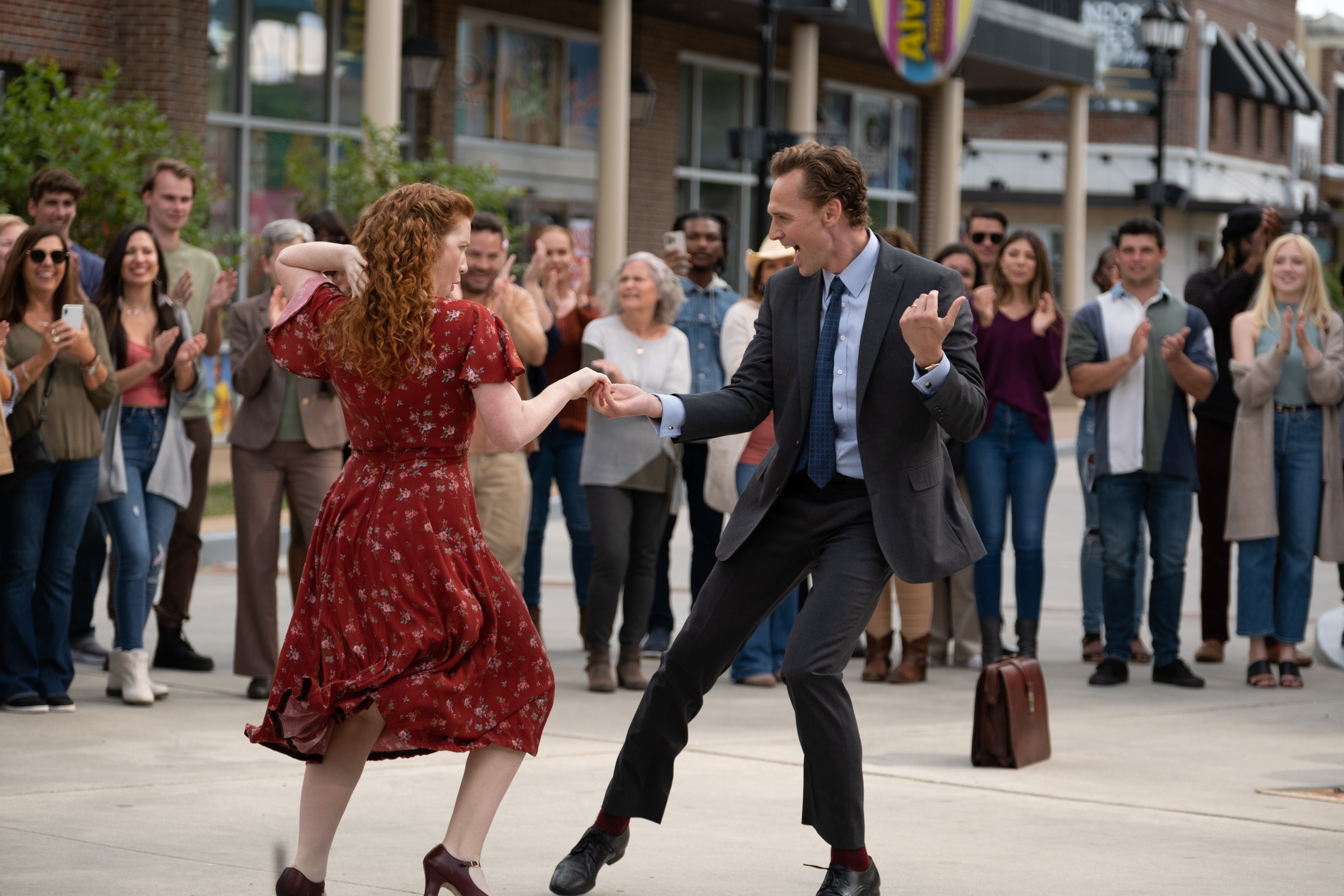 A red-haired woman (Annalise Basso) and a man in a suit (Tom Hiddleston as Charles Krantz) dance together in front of a crowd of onlookers at a mall complex. A film still from 'The Life of Chuck.'