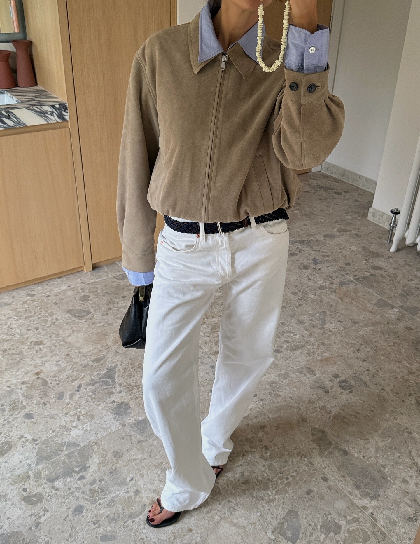 A woman taking a mirror selfie wearing white jeans, a suede jacket and open toe heels with a black pedicure