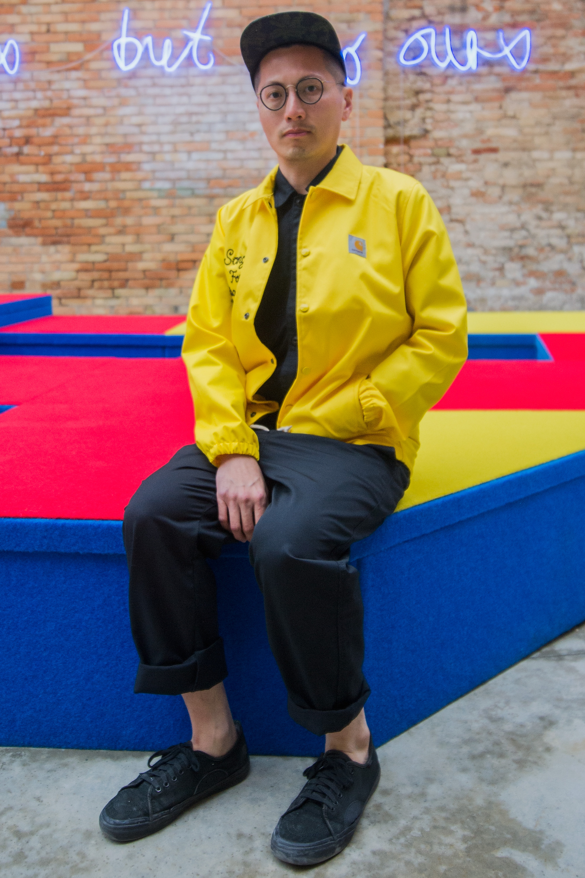 Multi-disciplinary artist Samson Young sits on a colorful bench wearing a black pants and hat and bright yellow jacket