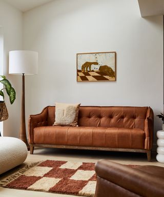 A white living room with a tan couch with a brown wall art print above it, a tall white lamp, and a light and dark brown checked rug
