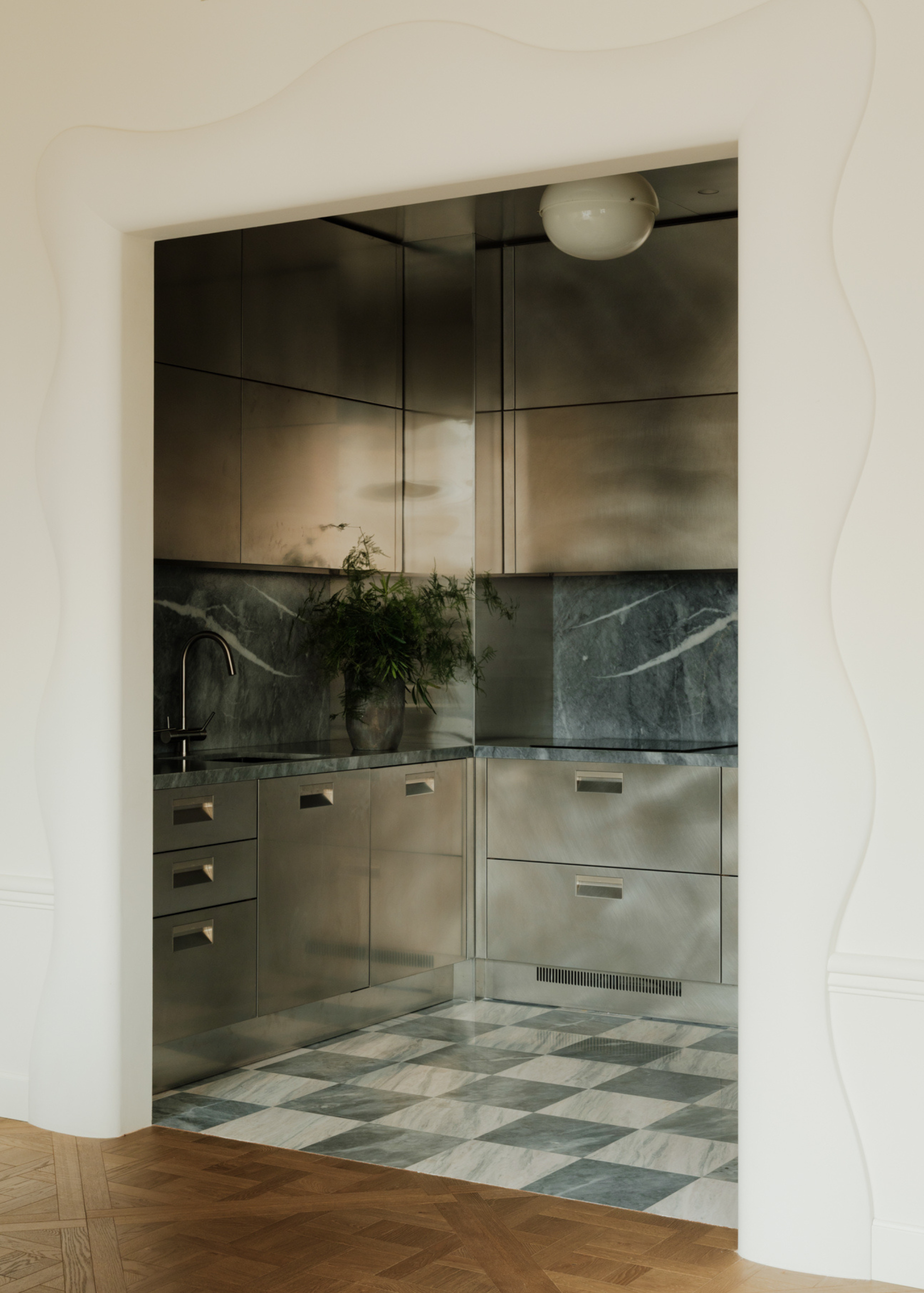 A kitchen with blue and white checkerboard marble flooring, brushed steel cabinets and drawers with a blue marble backsplash, and a vase of stems. Leading into the kitchen is a white wall with a 3D waved border