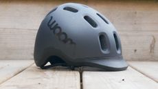 Side on view of the Woom Ready Kids Bike Helmet in black against a backdrop of a wooden deck