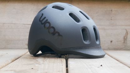 Side on view of the Woom Ready Kids Bike Helmet in black against a backdrop of a wooden deck