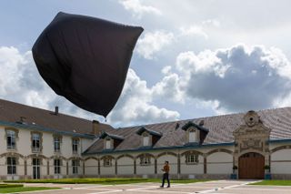 Daytime image, aeroglyphic inflated grey sculpture being flown by Tomás Saraceno on the ground attached to rope cords, courtyard type space, paved and grass areas,, surrounding white building, brown tiled roof, windows, large wooden arched door, blue cloudy sky