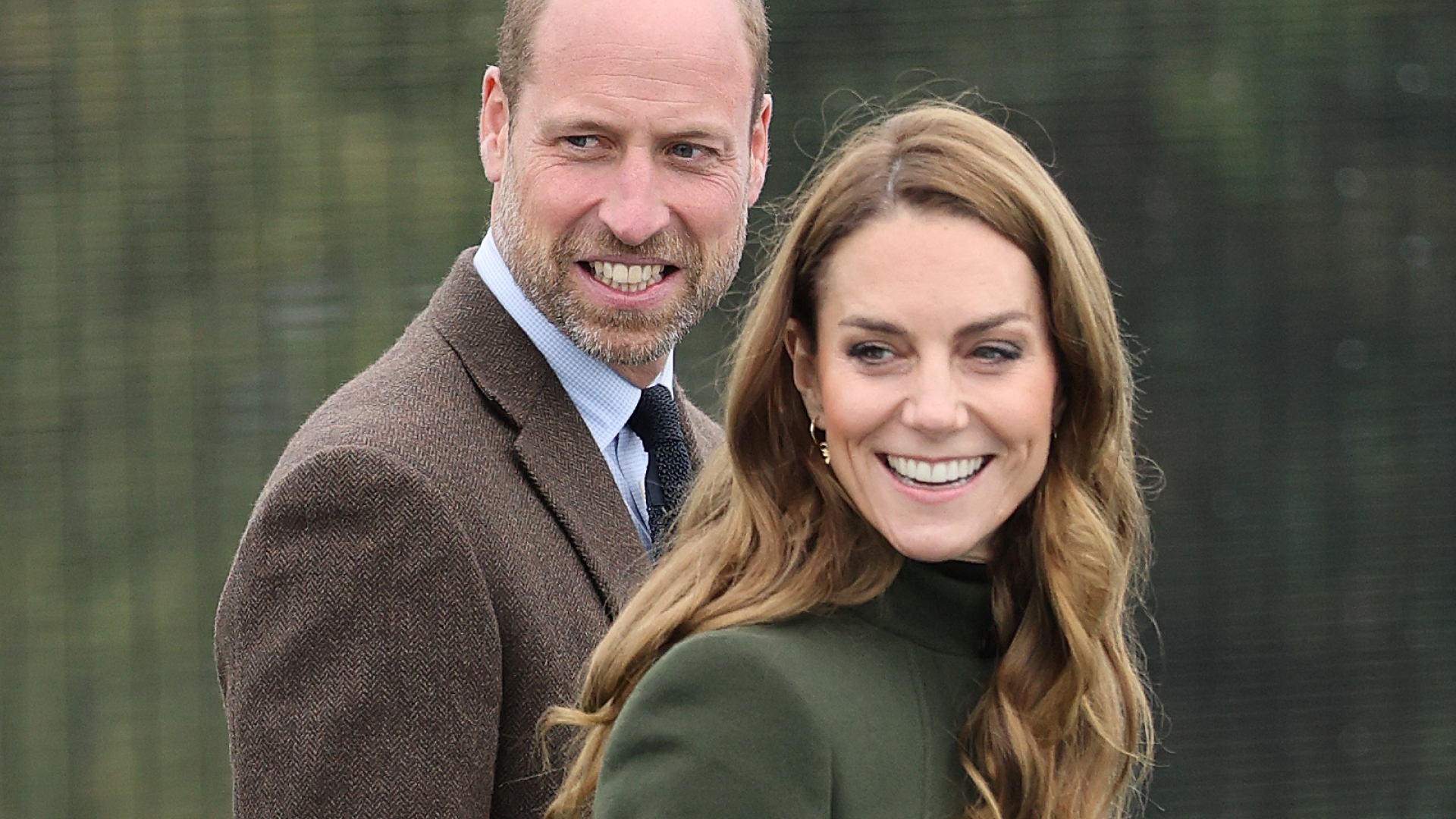 Prince William smiles while wearing a brown suit jacket and wife Kate Middleton wears a long green coat and has curly brown hair