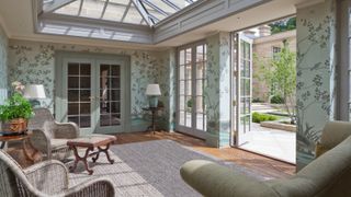 inside of orangery style sunroom with green floral wallpaper, wooden floor, patio doors to outdoors and connecting room, variety of furniture 