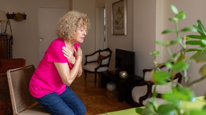Woman lowering herself into a chair in a domestic setting with her arms in an X-shape across her chest