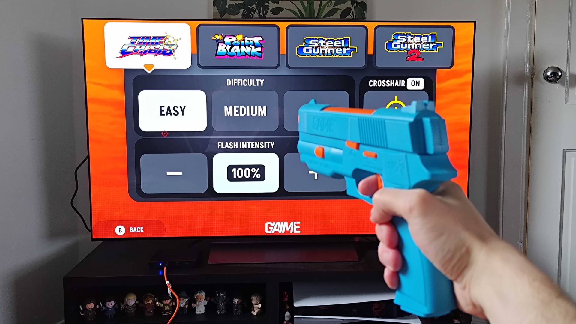 G&#039;AIM&#039;E Time Crisis Lightgun console connected to TV with hand using lightgun to navigate options menu.