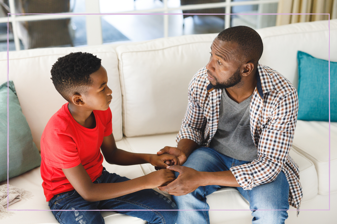 Serious african american father and son sitting on couch in living room talking and holding hands