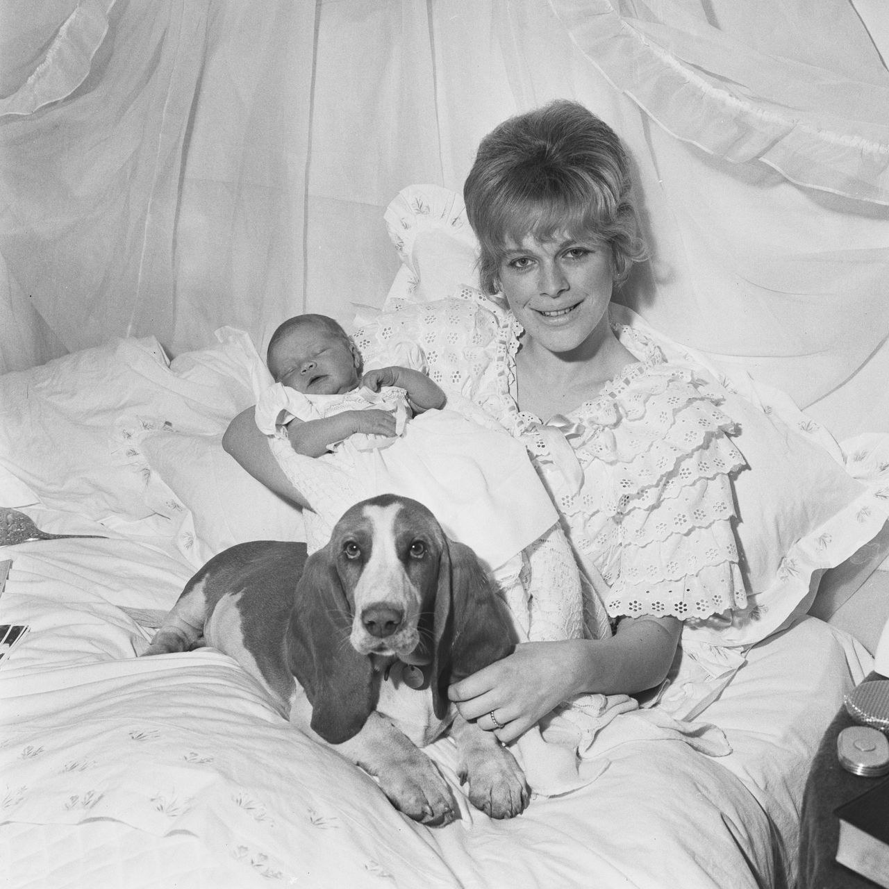 A woman sits up in bed holding a sleeping newborn while a basset hound lies contentedly beside them on the covers.