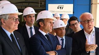 France's President Emmanuel Macron wearing a hard hat bearing the letters ST, is flanked by French government officials while the words ST Microelectronics can be seen on a wall behind them