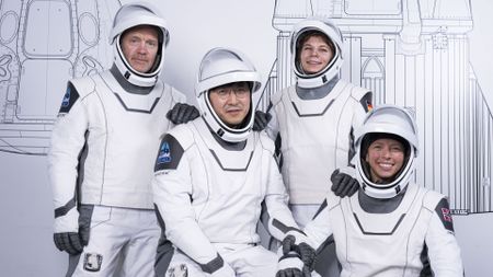 Four humans in white space suits post in front of a white wall with line drawings of a spaceship.